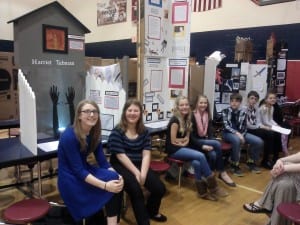 Six students sit in a line in front of three exhibit boards in a school cafeteria.